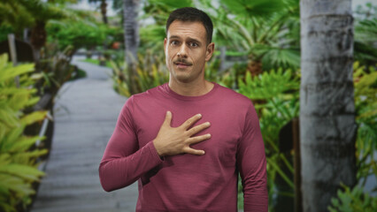 Fototapeta premium Hispanic man with mustache, hand on chest on boardwalk path, wearing maroon long sleeve shirt and looking earnest; sincerity gratitude.