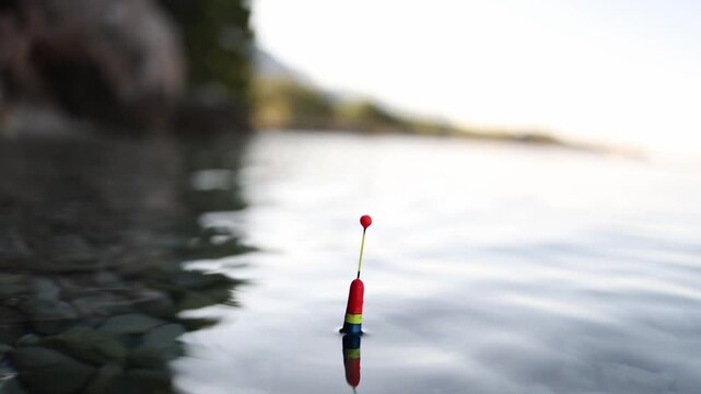 Fishing float rests quietly on clear sea water above visible stones moving gently with small waves while the angler waits patiently for any sign of a fish near shore here