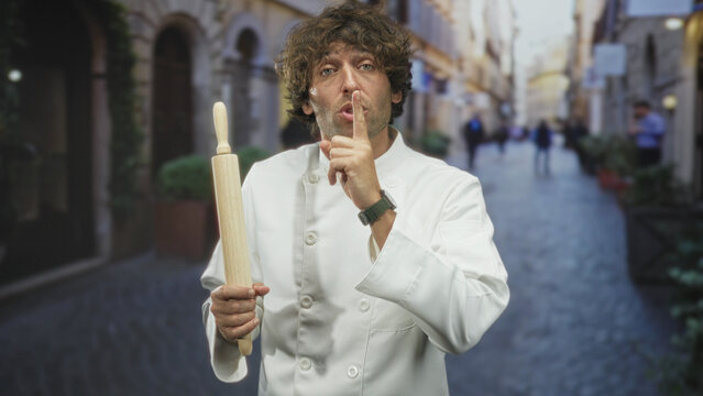 Man chef in white uniform holds a rolling pin and places finger to lips for silence on a narrow cobblestone street with blurred passersby; playful secrecy.