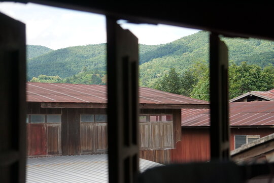 framed view through open wooden window shutters looking out at traditional wooden houses with tin roofs set against a lush, green mountain range under a cloudy sky. The composition creates a peaceful,