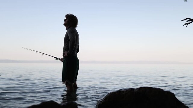 Disappointed fisherman stands knee deep in the sea with his rod lowered as another fruitless effort brings no bite leaving him frustrated after a long wait near the shore