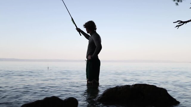 Fisherman slowly works his pole in shallow coastal water checking the line and tackle while enjoying a quiet evening outdoors and searching for fish close to shore nearby
