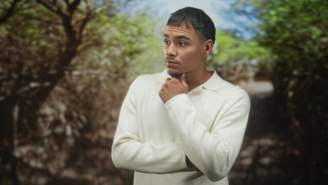 Man touching chin with hand in a pensive thinking pose on a forest path, wearing white sweater; thoughtful reflection.
