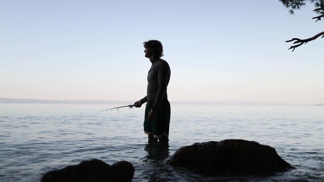 Male angler stands in clear shallow sea water and carefully controls his fishing rod while watching the surface for movement during a relaxed coastal fishing trip at dusk