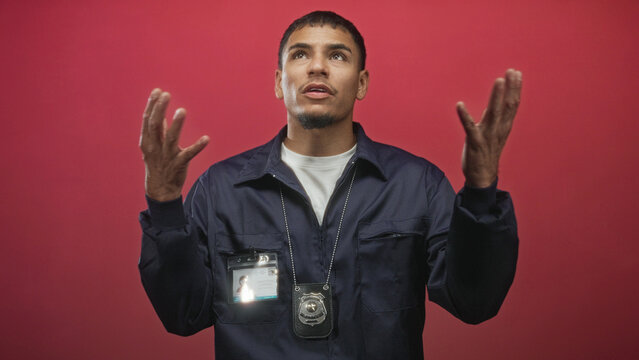 Young hispanic man raises hands showing police badge and idcard against red backdrop in studio; contemplation.