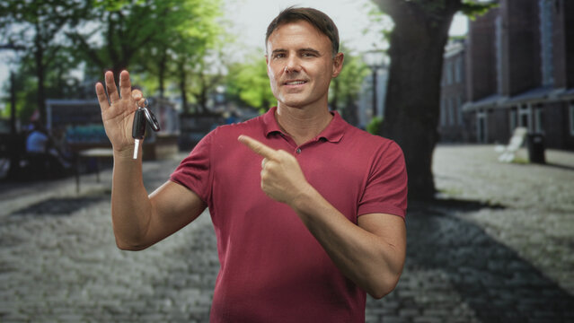 Man holding car keys and pointing index finger on cobbled street plaza with trees and benches in front of brick buildings wearing red polo shirt; confidence trust.