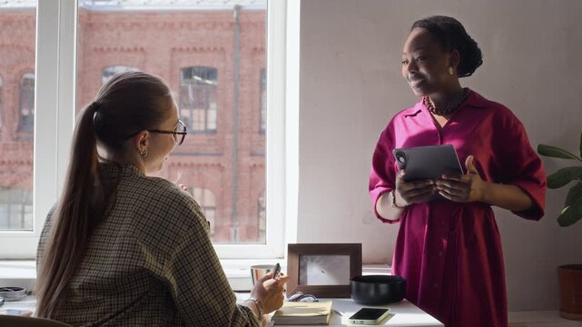 African American young adult woman holding tablet and discussing project with Caucasian colleague sitting at desk. Video is perfect for corporate training and diversity in workplace marketing