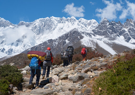 Group of porters and hikers with heavy backpacks walking up rocky mountain trail towards majestic snow peaks in Nepal Himalayas.