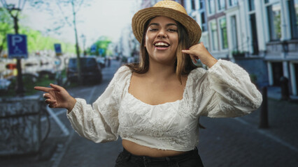 Fototapeta premium Woman in straw hat and white embroidered crop top makes call me hand gesture and points finger on cobblestone street; playful.