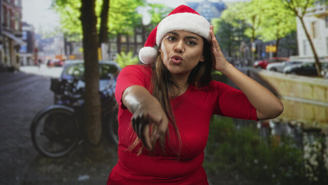 Woman wearing santa hat points hand to camera on street, puckering lips and adjusting hat with other hand; festive joy.