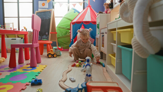 Girl kneeling on foam mat playing with wooden train track and toy car, hand guiding cars beside robot and bowling pins in a colorful playroom building; curiosity imagination.