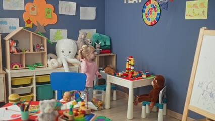 Fototapeta premium Little blonde girl stacks colorful blocks at a plastic play table amid plush toys and shelving in a preschool building; curiosity wonder imagination play.