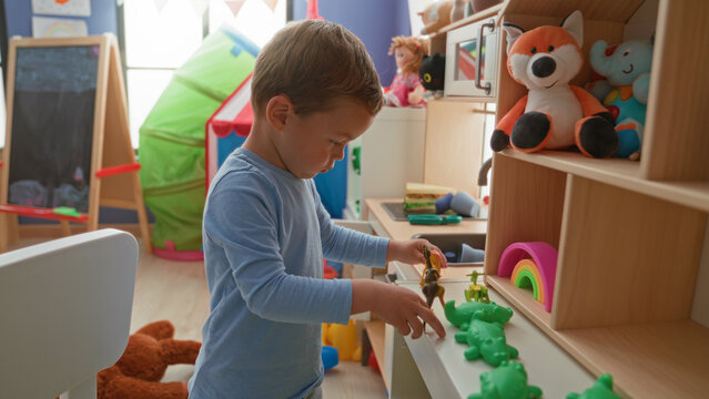 Boy holding green toy dinosaur with bare hands while arranging plastic frogs on a low shelf in a playroom building; curiosity learning.
