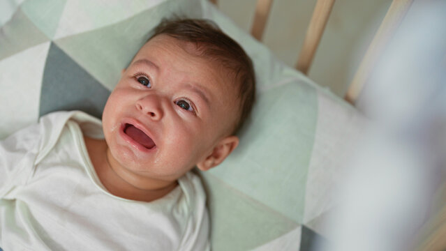 Hispanic baby boy holds wooden teether to mouth while lying on a patterned crib sheet inside a building crib; curiosity teething comfort.