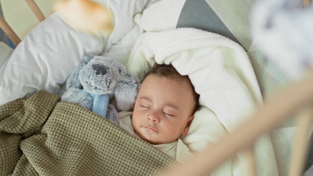 Hispanic baby sleeping with closed eyes and blanket, stuffed toy visible in crib inside building; calm rest.