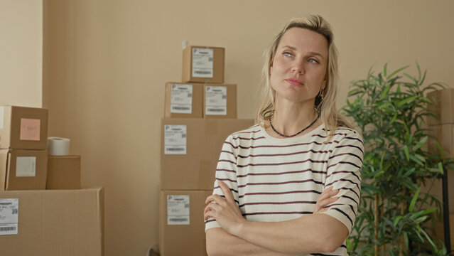 Blonde woman with stacked cardboard packages, arms crossed and looking up with pensive expression in a building; small business determination.