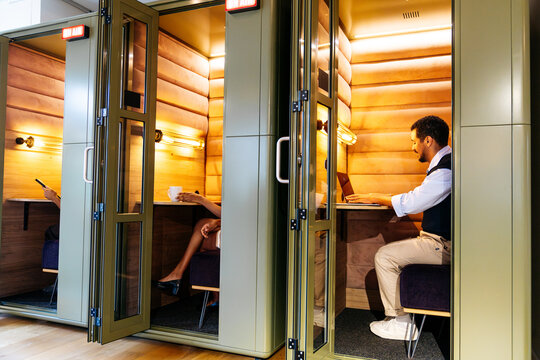 People working and communicating in private soundproof booths within a modern collaborative office space, focusing on tasks