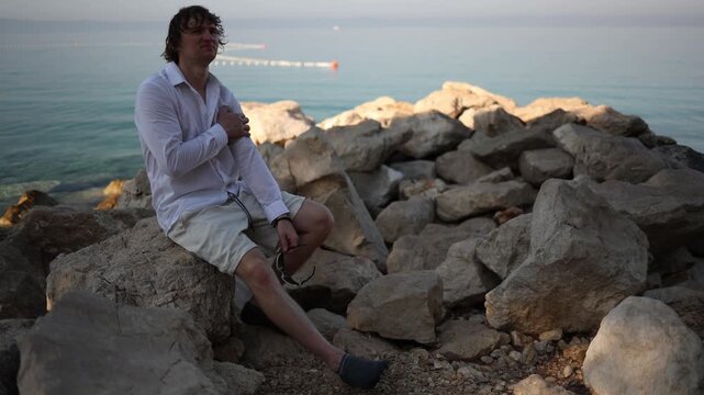 Young man feels uncomfortable relaxing by the sea after constant work sitting awkwardly on coastal rocks as he struggles to switch off and find what to do during vacation

