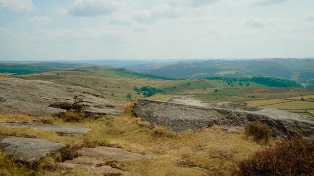 Flat gritstone rock platform above open countryside and rolling hills, slide shot. Heather, dry grass and weathered stone under soft cloudy sky. Elevated moorland landscape with agricultural fields