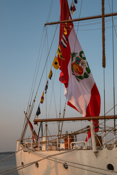 Sailing on a tall ship with flags and a large Peruvian flag at sunset. A tall ship sails during sunset, displaying a large Peruvian flag. Colorful flags are seen along the mast.