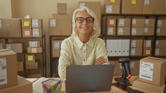Woman at desk with laptop and arms crossed in a small business building amid stacked cardboard boxes and barcode scanner; entrepreneurship confidence.