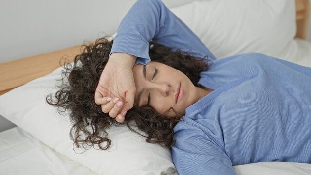 Young woman with hand on forehead resting on bed in bedroom, eyes closed and curly hair on pillow, wearing blue pajamas; fatigue rest.