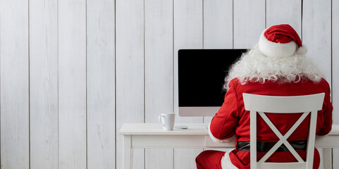Obraz premium Santa Claus in red suit sitting on a white chair looking at a monitor screen,