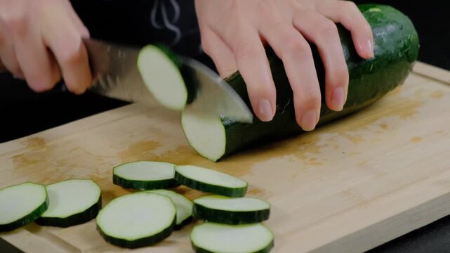 Hands in black clothing slice a dark green zucchini with a large knife on a wooden cutting board during daytime in a Cooking kitchen, close-up view.