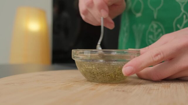 Hands in green leaf-patterned apron stir thick greenish-yellow herb mixture in glass bowl on wooden counter, Cooking location, daytime.