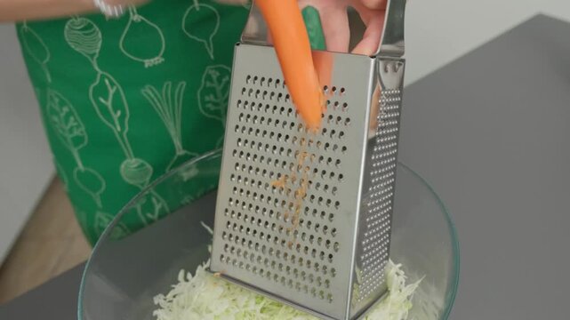 Close-up of hands grating a carrot into a bowl of shredded cabbage in a kitchen, cooking preparation, daytime.