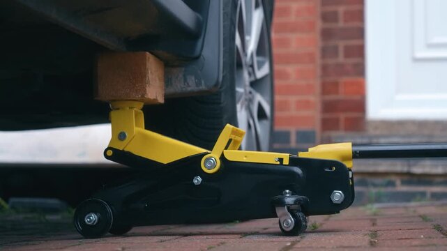 Raising vehicle with hydraulic floor jack and wooden spacer during car maintenance procedure. Automotive repair concept with controlled lifting on driveway for wheel access and underbody work. DIY