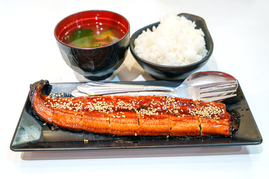 Grilled unagi kabayaki with sesame seeds, served with a bowl of steamed white rice and miso soup. Authentic Japanese eel set meal with cutlery on white background. Healthy, savory gourmet seafood.