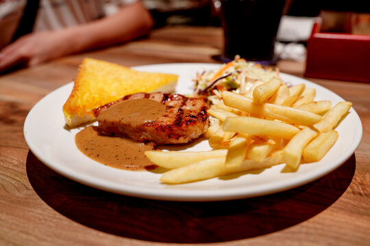 Grilled pork chop topped with savory brown gravy served with crispy golden French fries, buttered toast, and fresh coleslaw on a white plate. Gourmet meal in a restaurant setting on wood table.