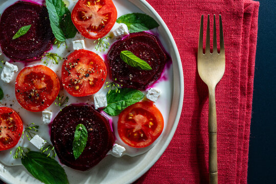 Gourmet beetroot and tomato salad, closeup. Beautiful flat lay of fresh summer salad with sliced red beets, tomatoes, feta cheese cubes, and mint on white yogurt sauce base