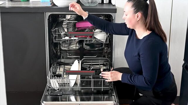 Footage of a young woman loading plates into an open dishwasher in a modern home kitchen reaching into the rack a candid real-life lifestyle scene of daily housework and cleaning up at home.