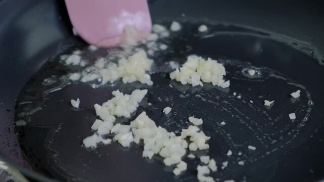 Close-up of stirring chopped garlic or shallots in oil with pink spatula in dark pot on stovetop, Cooking.