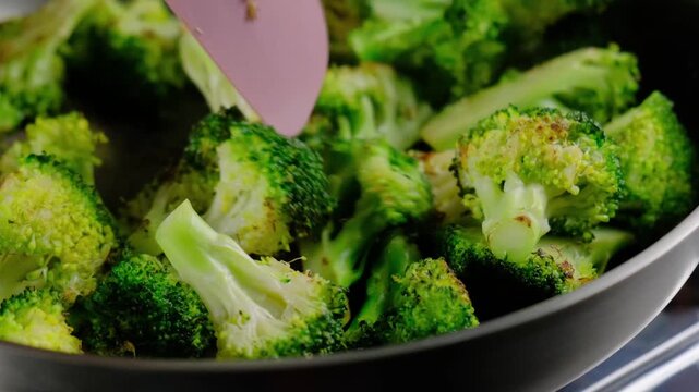 Close-up of broccoli florets and stems being stir-fried in a dark pan with a pink spatula, showing caramelization, in a bright indoor kitchen at the Cooking location.