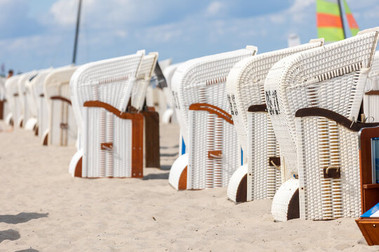 Rows of white wicker beach chairs line sandy Kuhlungsborn Baltic coast bright summer sky. Colorful sail, soft shadows and repeating patterns create relaxed seaside vacation mood with coastal charm