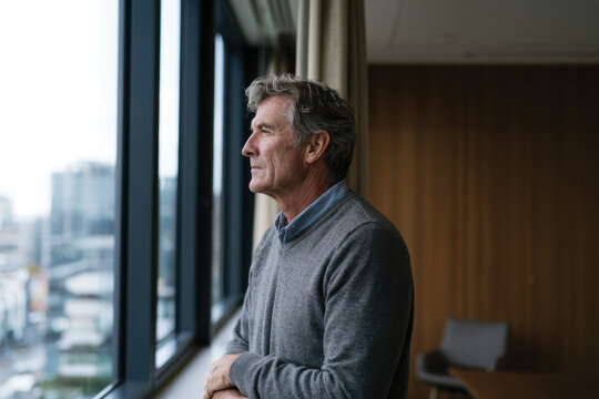 A mature man stands by the window in a contemporary office, gazing out thoughtfully, capturing a moment of reflection amid the vibrant energy of the city outside.