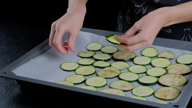 Close-up of hands in dark patterned clothing slicing zucchini and eggplant on a parchment-lined baking sheet during food preparation in a Cooking kitchen.