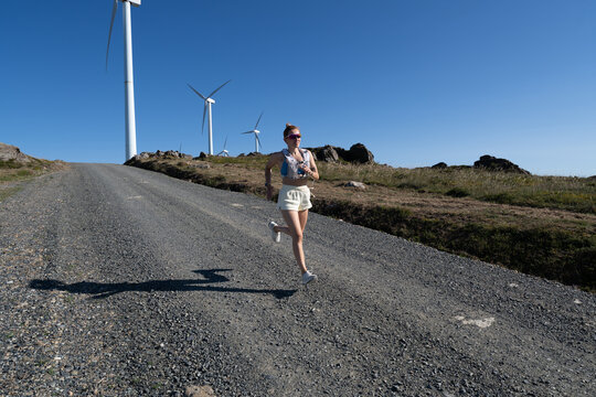 A white woman jogging on a gravel road near wind turbines in Galicia