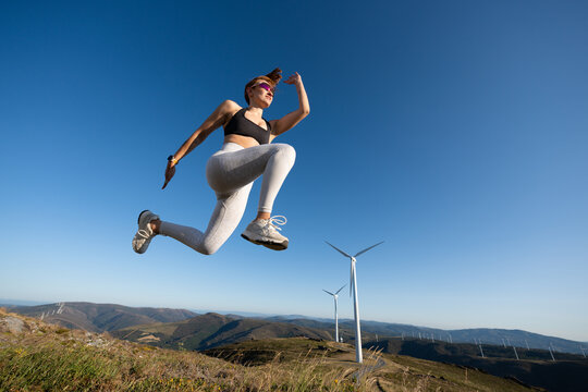 A white woman jumps on a hilltop near wind turbines in Galicia, Spain