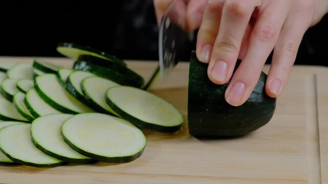 Close-up of hands slicing a dark green zucchini with a serrated knife on a wooden board in a Cooking setting, slices arranged nearby, studio lighting, daytime.