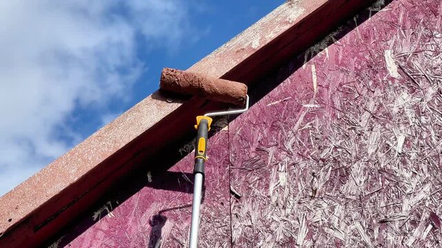 Low-angle view of painting the wooden fascia trim of a shed roof using a roller pole with brown paint against a blue sky with clouds during home renovation.