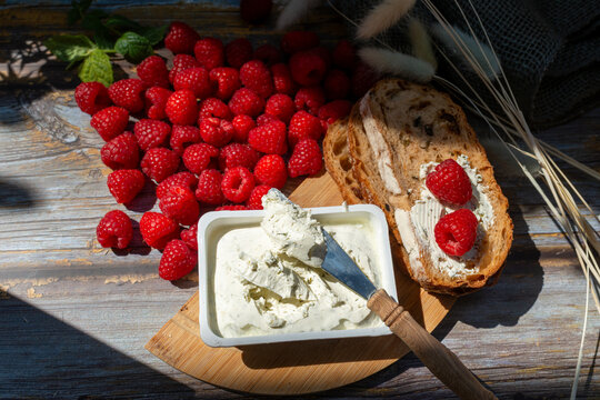 Artisan bread spread with herb cream cheese and topped with ripe raspberries Rubus idaeus, served beside an open cheese container and wooden-handled knife. Creative sweet and savory breakfast or summe