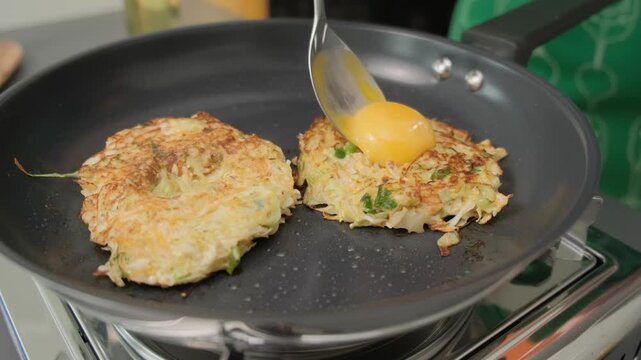 Cooking vegetable fritters in a frying pan, pouring raw egg yolk over them, in a kitchen setting at Cooking location.