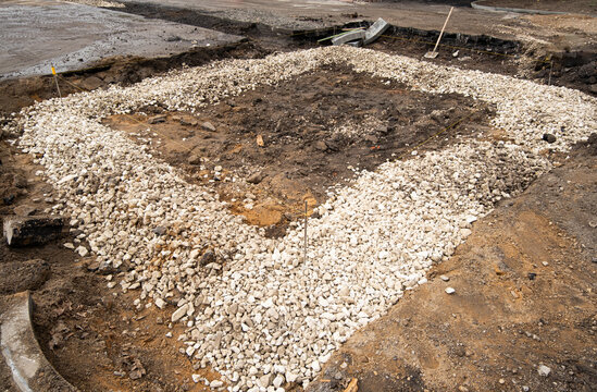 Workers prepare the foundation at a construction site. Gravel is placed around the area while soil is visible in the center. Tools are scattered nearby.