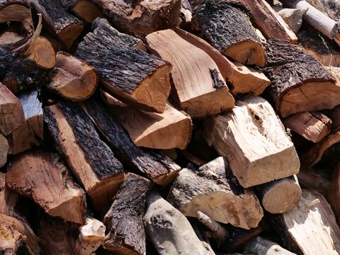 Stack of Cut and Split Firewood - A close-up photograph of a large, irregularly stacked pile of cut and split firewood logs. The logs show various textures of bark and the grain 