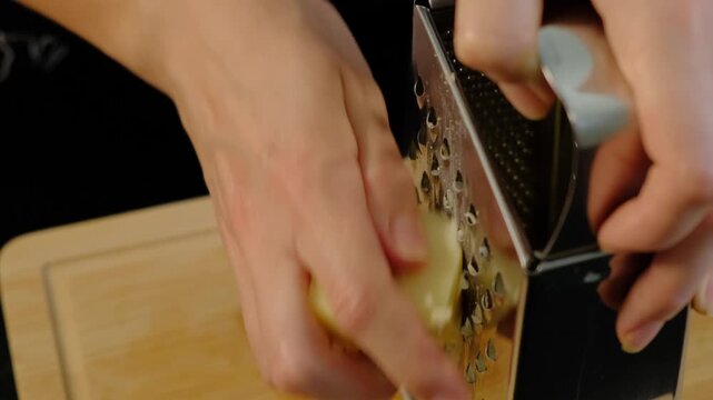 Close-up of adult hands grating a light yellow cheese on a wooden cutting board in a kitchen during daytime cooking.