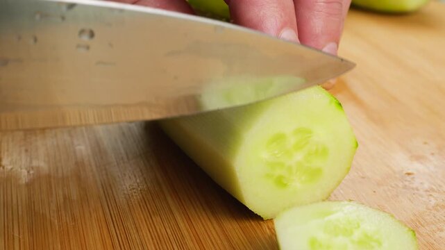 A chef uses a sharp knife to slice a fresh green cucumber into round pieces on a wooden board, neatly stacking the clean organic slices for professional cooking preparations.